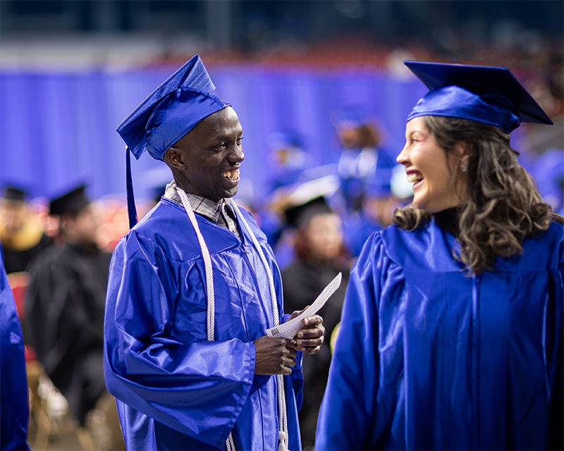 Two Madison College graduates wear blue caps and gowns and smile broadly at each other. 