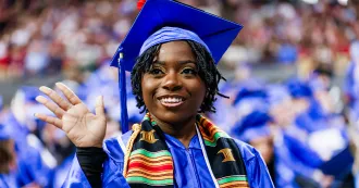 Black student grad, wearing blue cap and gown, waving at friend at 2025 Spring commencement ceremony.