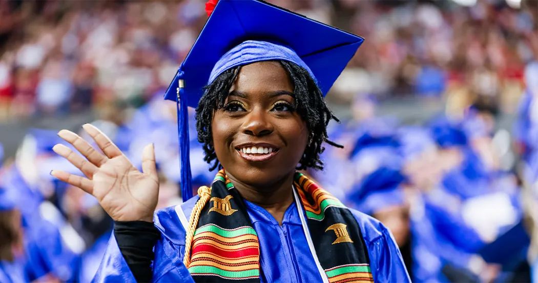 Black student grad, wearing blue cap and gown, waving at friend at 2025 Spring commencement ceremony.