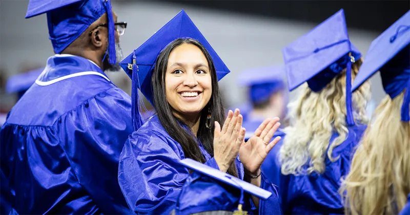 Madison College graduate smiling and clapping wearing blue cap and gown