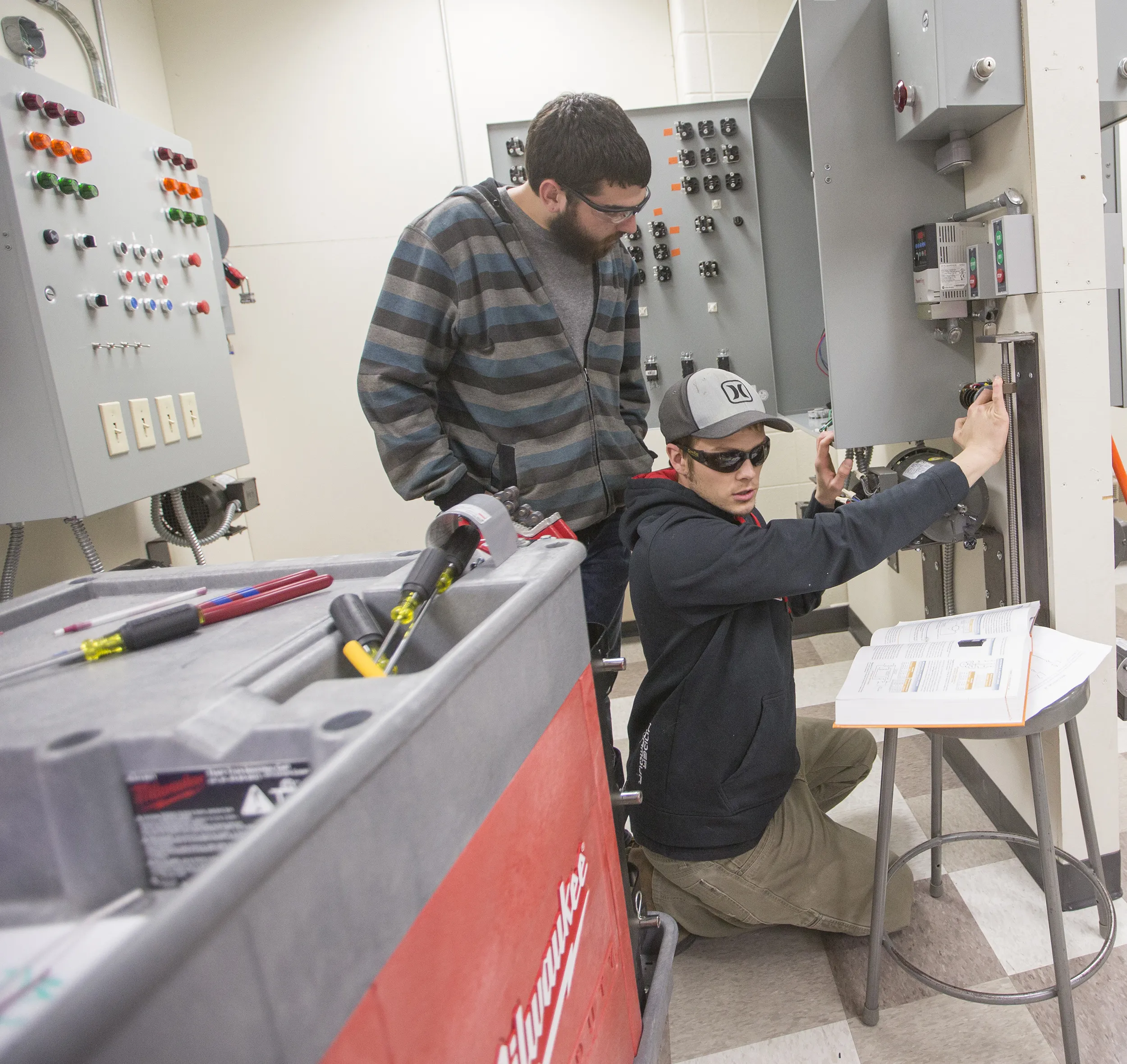 Two male students, both with dark hair and glasses, work with electrical boxes in a classroom.