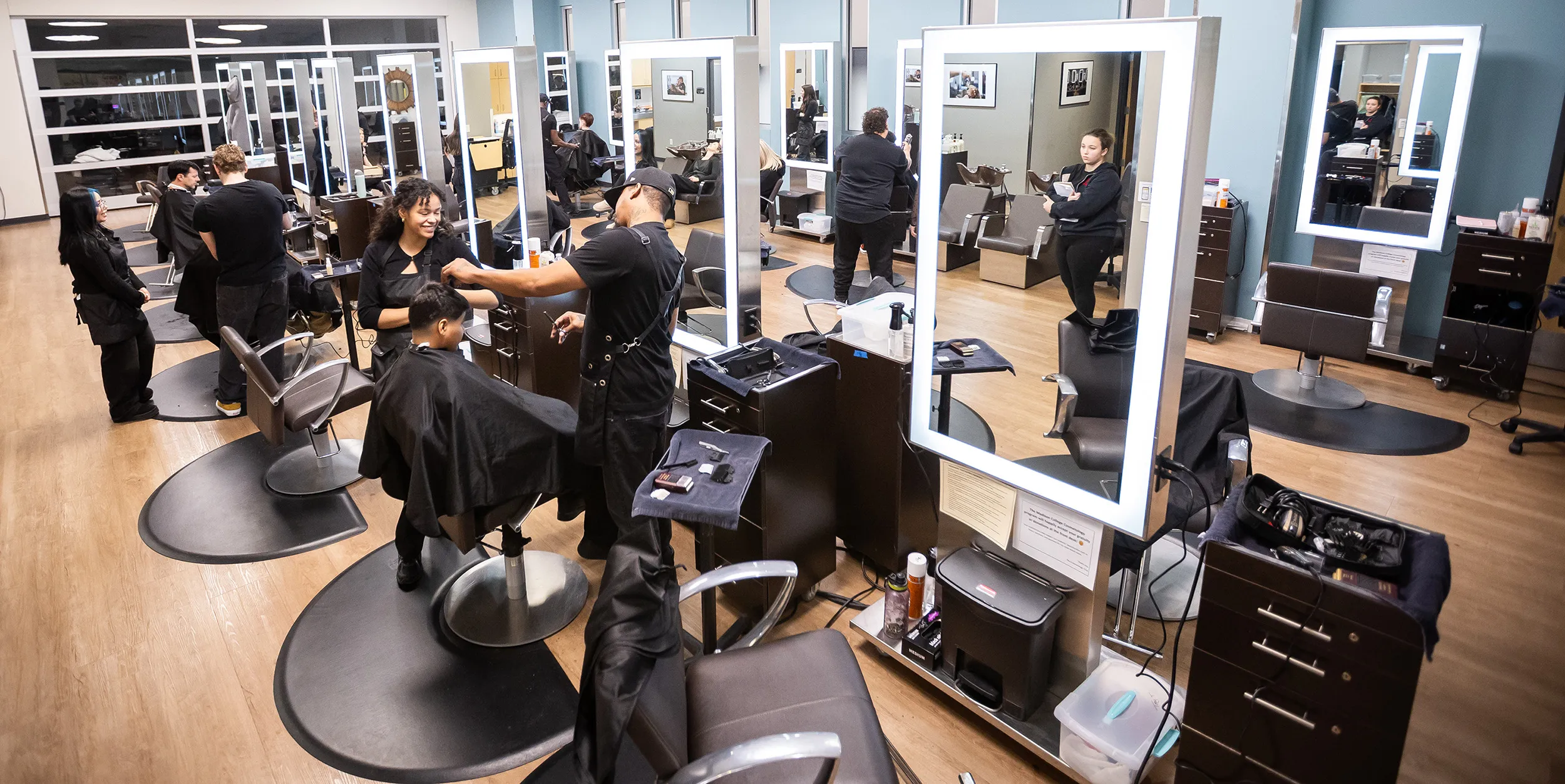 Barbering program classroom with client chairs, mirrors and equipment cabinets. Students in foreground cutting client's hair.