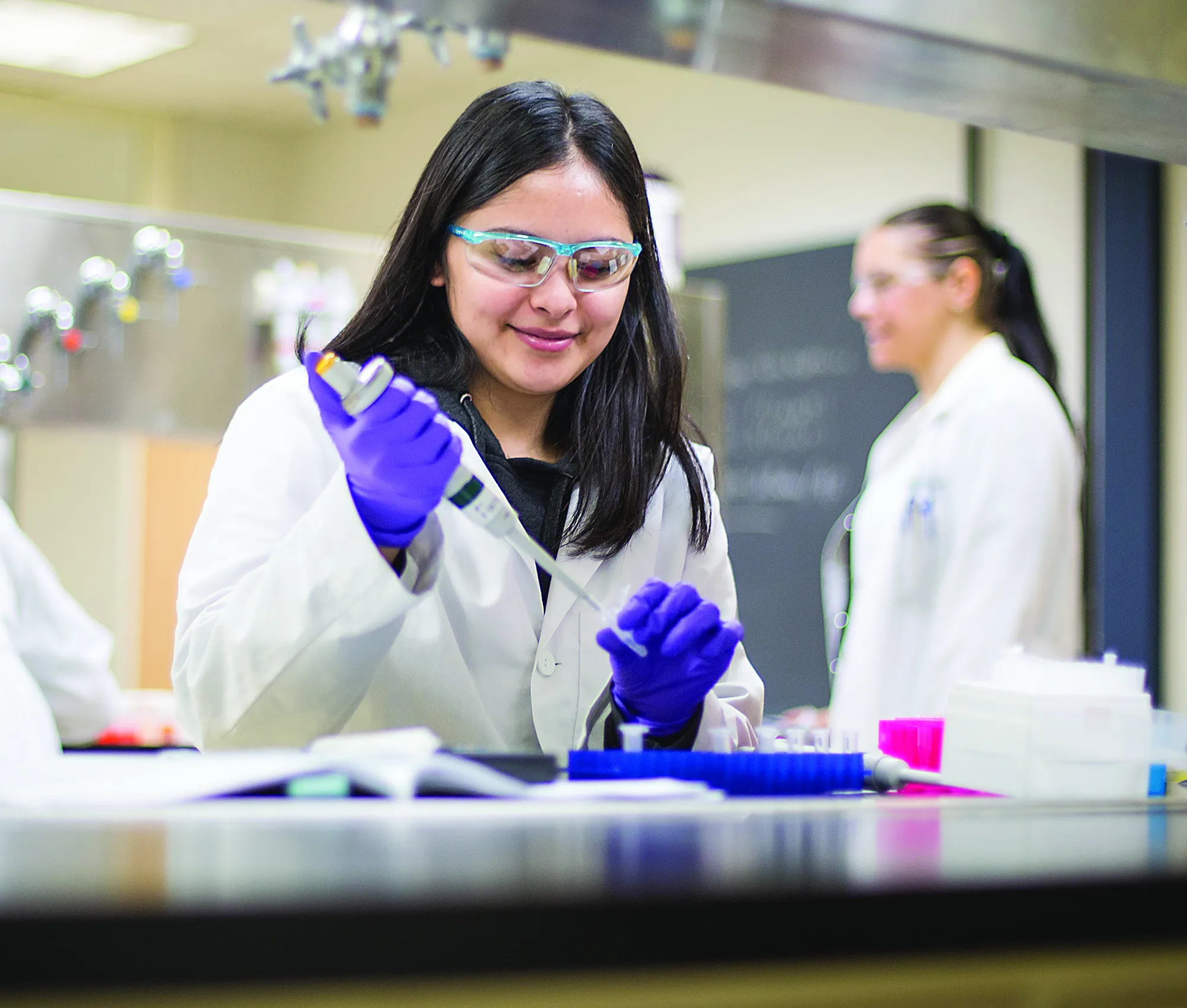 Students with black hair, wearing a white lab coats, holding a syringe in a lab setting.