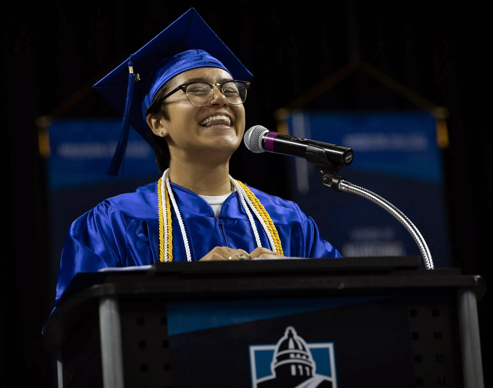 Grad with blue gown, dark glasses, dark hair, smiling, while standing at a podium.