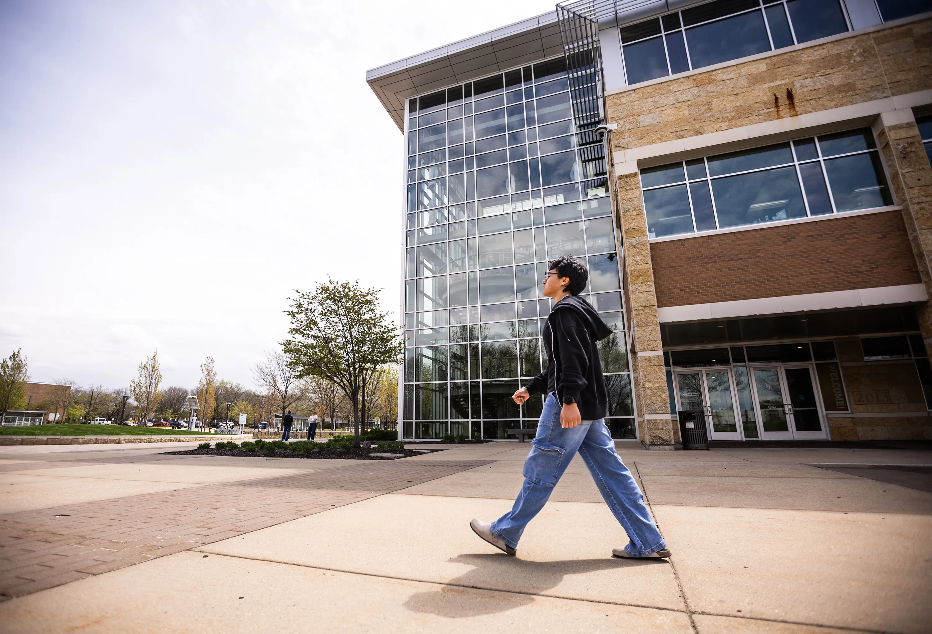 Student walking on campus, with building behind her, gray concrete and green trees.