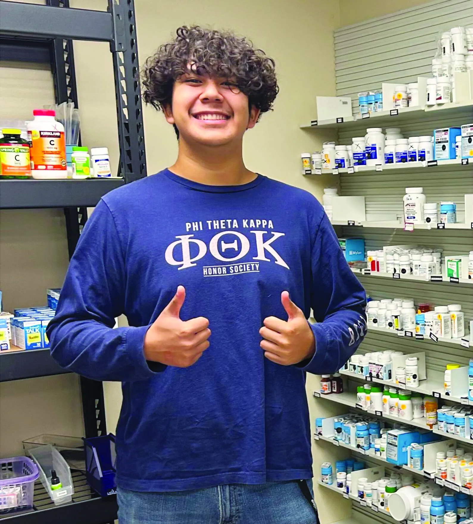 Student standing in a pharmacy room, wearing a blue shirt and giving the thumbs-up.