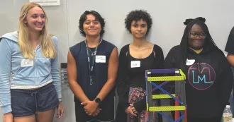 Photo of students standing in front of a classroom white board smiling at the camera.