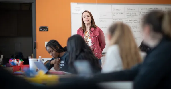 Instructor standing in room next to whiteboard talking while students listen in foreground.