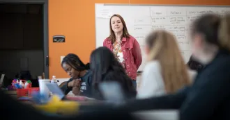 Instructor standing in room next to whiteboard talking while students listen in foreground.