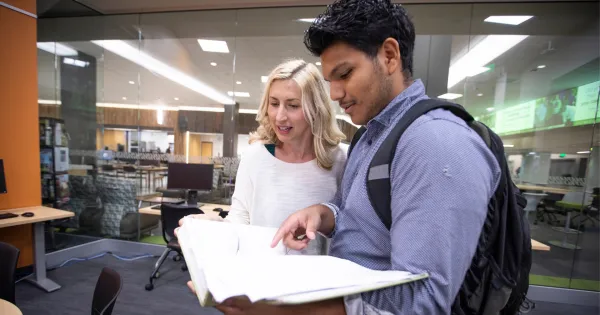 A female and male student looking at pages in a book, while talking in the library.