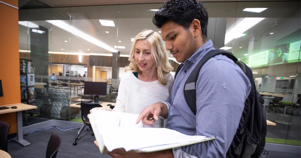 A female and male student looking at pages in a book, while talking in the library.