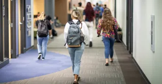 Three students, wearing blue jeans and carrying backpacks, walking in a hallway at the Madison College Truax campus.