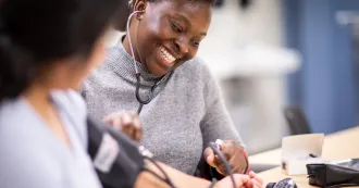 Woman nursing student putting a blood pressure cup around a patient student's arm and reading the device.
