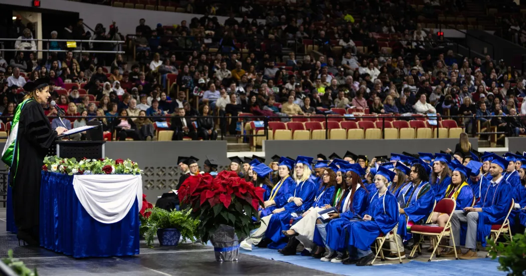 Graduates seated, wearing blue cap and gowns, President Berne at the podium speaking.