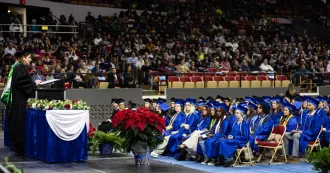 Graduates seated, wearing blue cap and gowns, President Berne at the podium speaking.