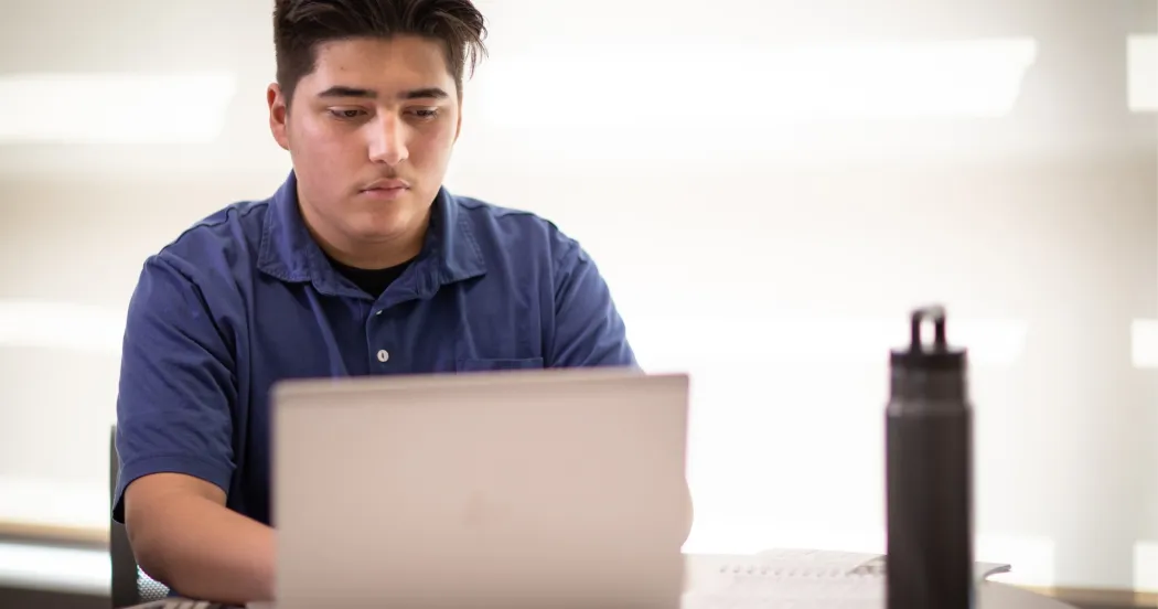 Student in blue shirt, working on a laptop, water bottle on desk.