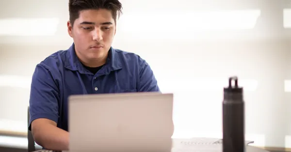 Student in blue shirt, working on a laptop, water bottle on desk.