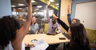 Students sitting around a table in library meeting space, holding hands up in celebration and laughing and smiling.