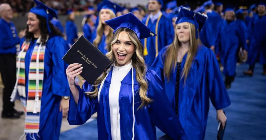 Student in blue cap and gown, smiling as she holding a Madison College diploma that she received at the Commencement ceremony.