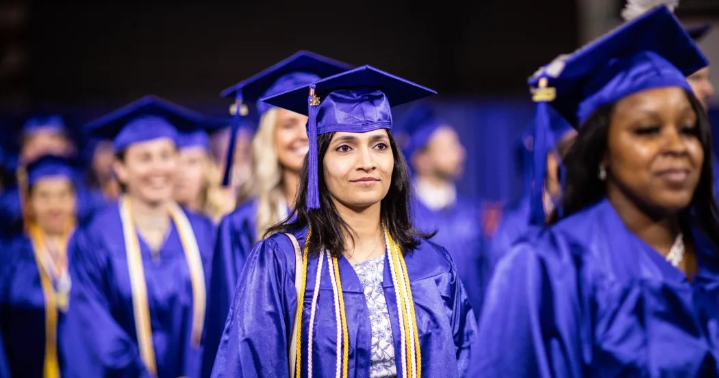 Female grad at commencement ceremony walking down aisle, wearing blue cap and gown and smiling.