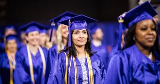 Female grad at commencement ceremony walking down aisle, wearing blue cap and gown and smiling.