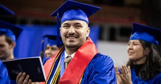 Photo of grad at fall 2025 commencement smiling, wearing blue cap and gown and holding a commencement program.
