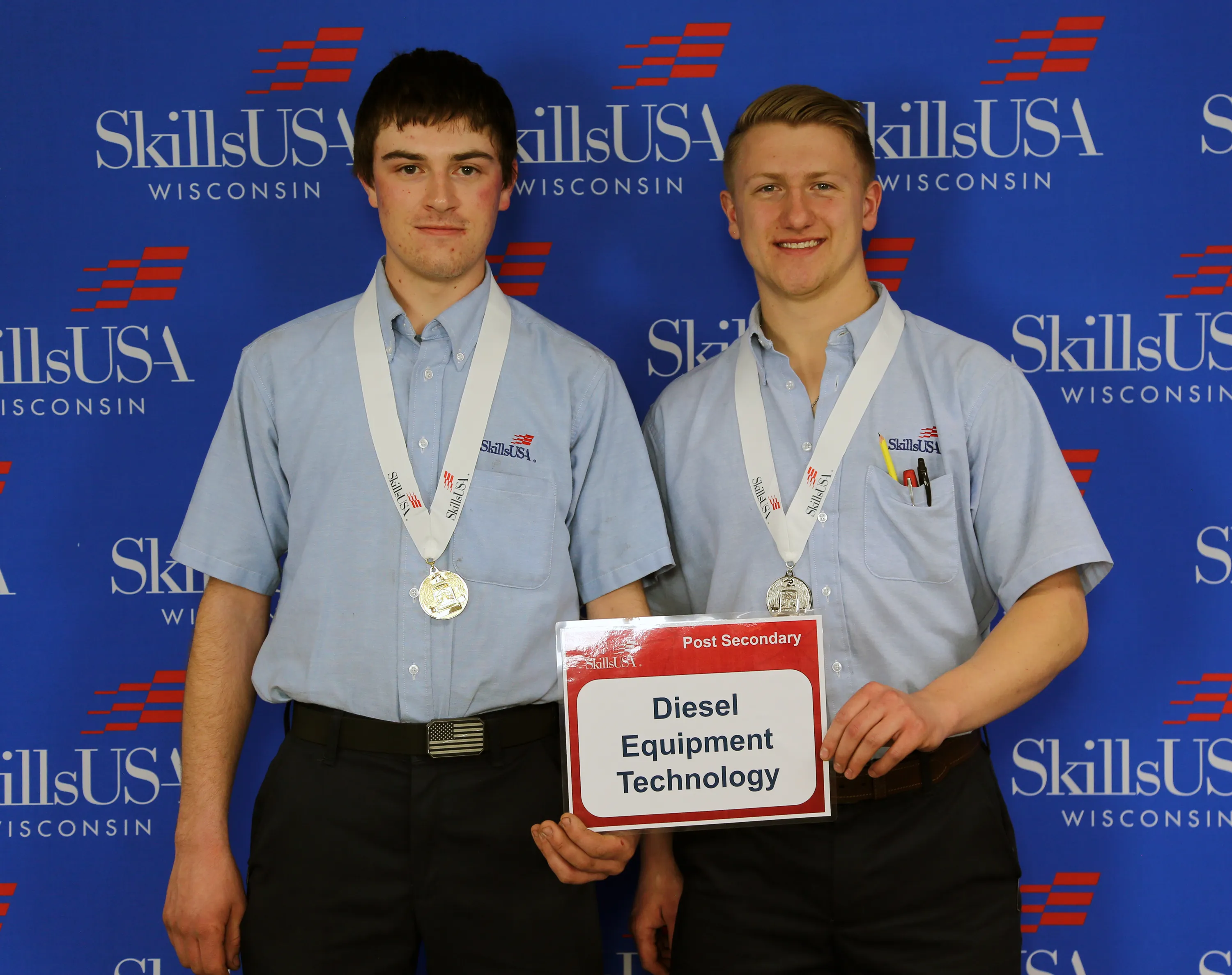 Two students, wearing light blue shirts and medals, standing in front of a blue SkillsUSA banner.