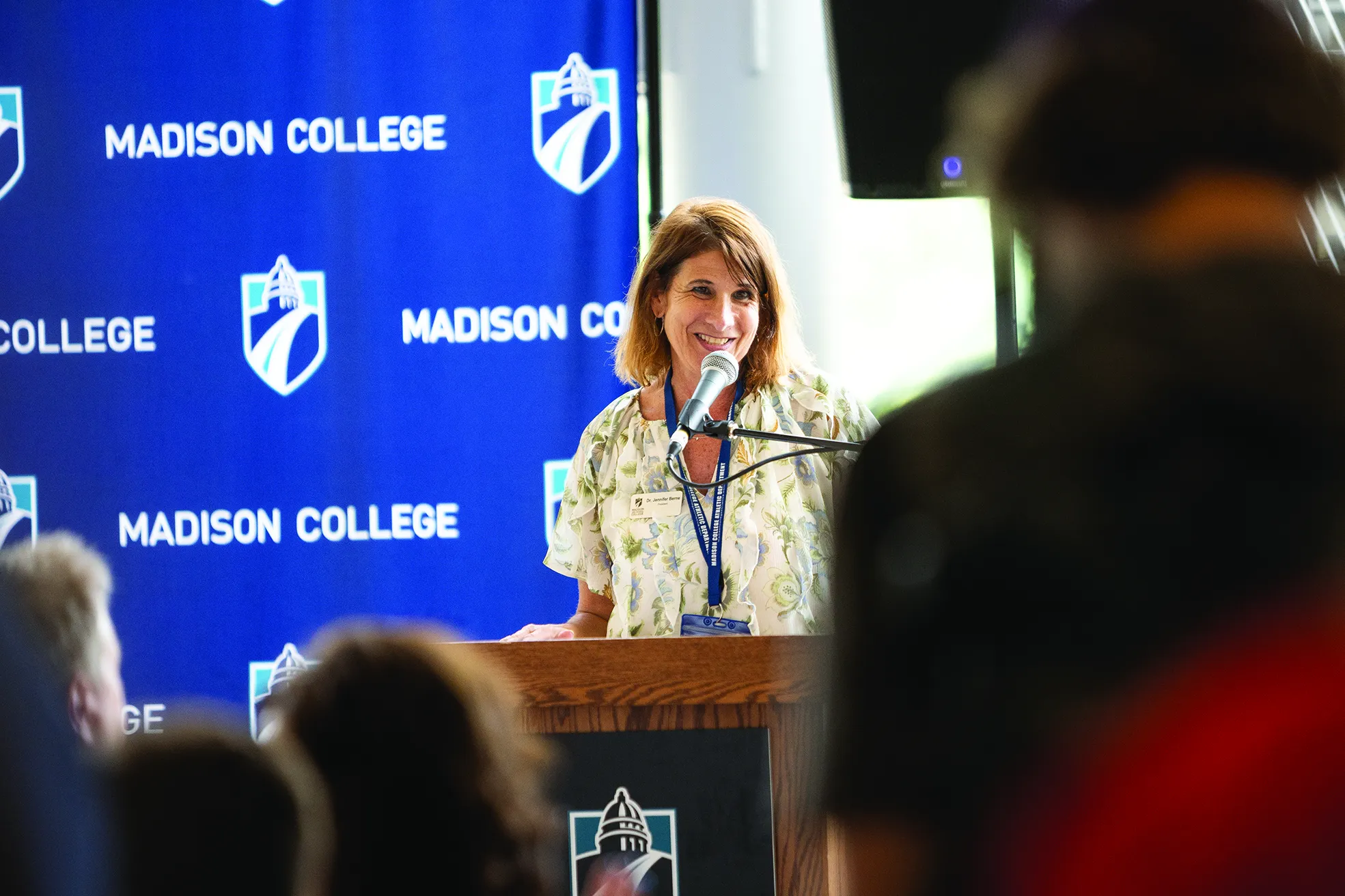 President Berne at a podium, speaking into a microphone, in front of a blue Madison College banner.