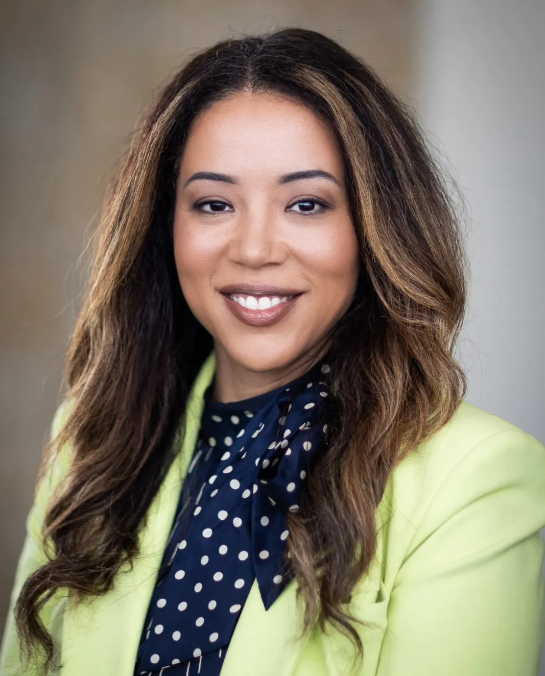 Woman with brown hair, blue blouse and yellow blazer, smiling.
