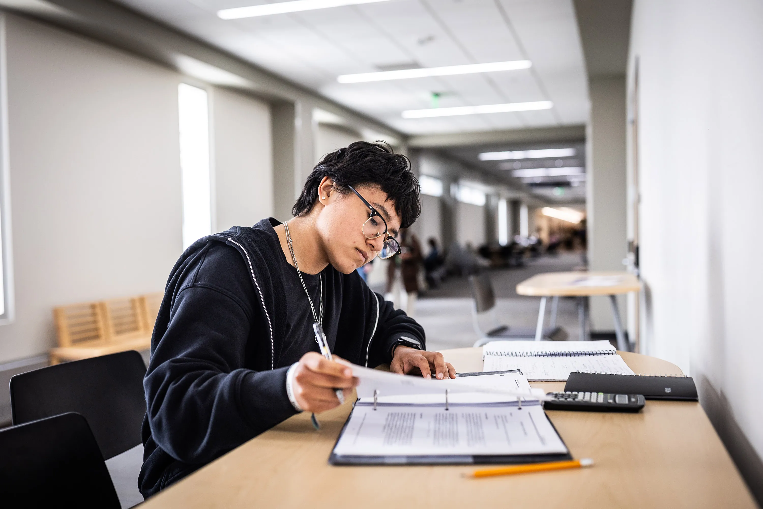 Student with dark hair and dark glasses, wearing a dark blue sweatshirt, looking at notebook on table.