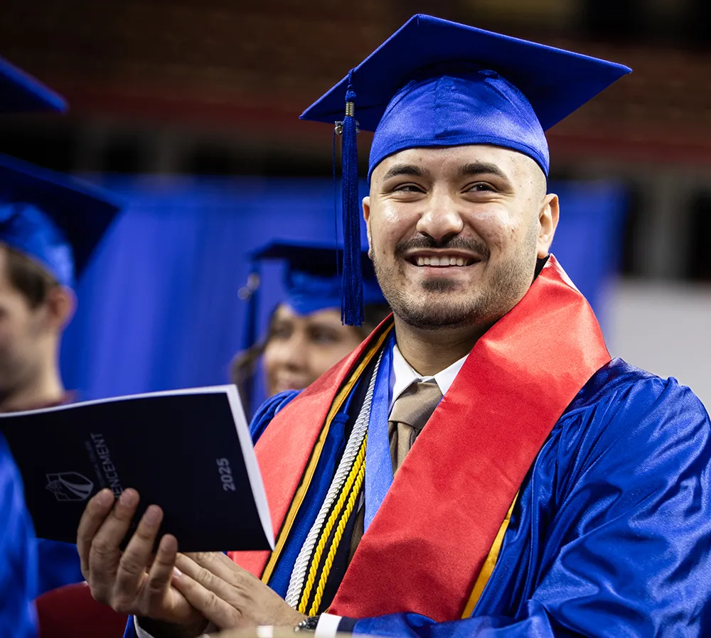 Male graduate wearing blue cap and gown with red sash, clapping and smiling.