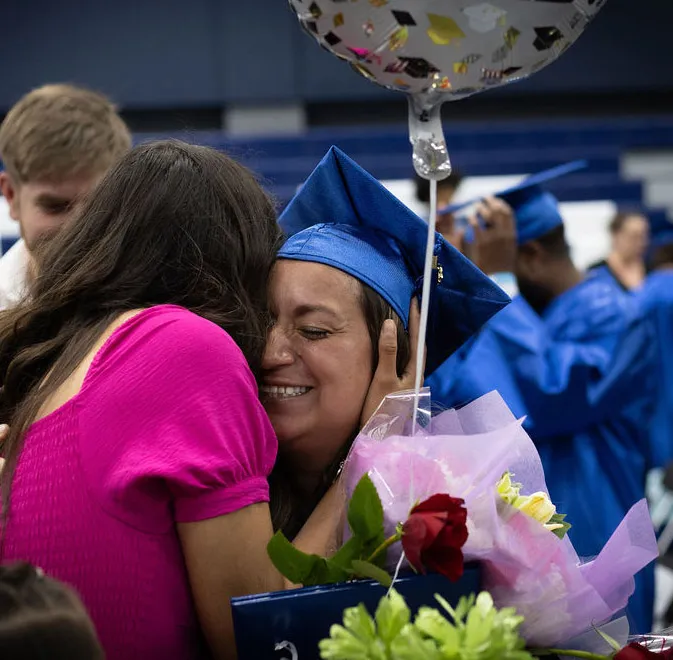 Woman with blue grad cap hugging a person. There is a red rose and silver balloon in her hand.