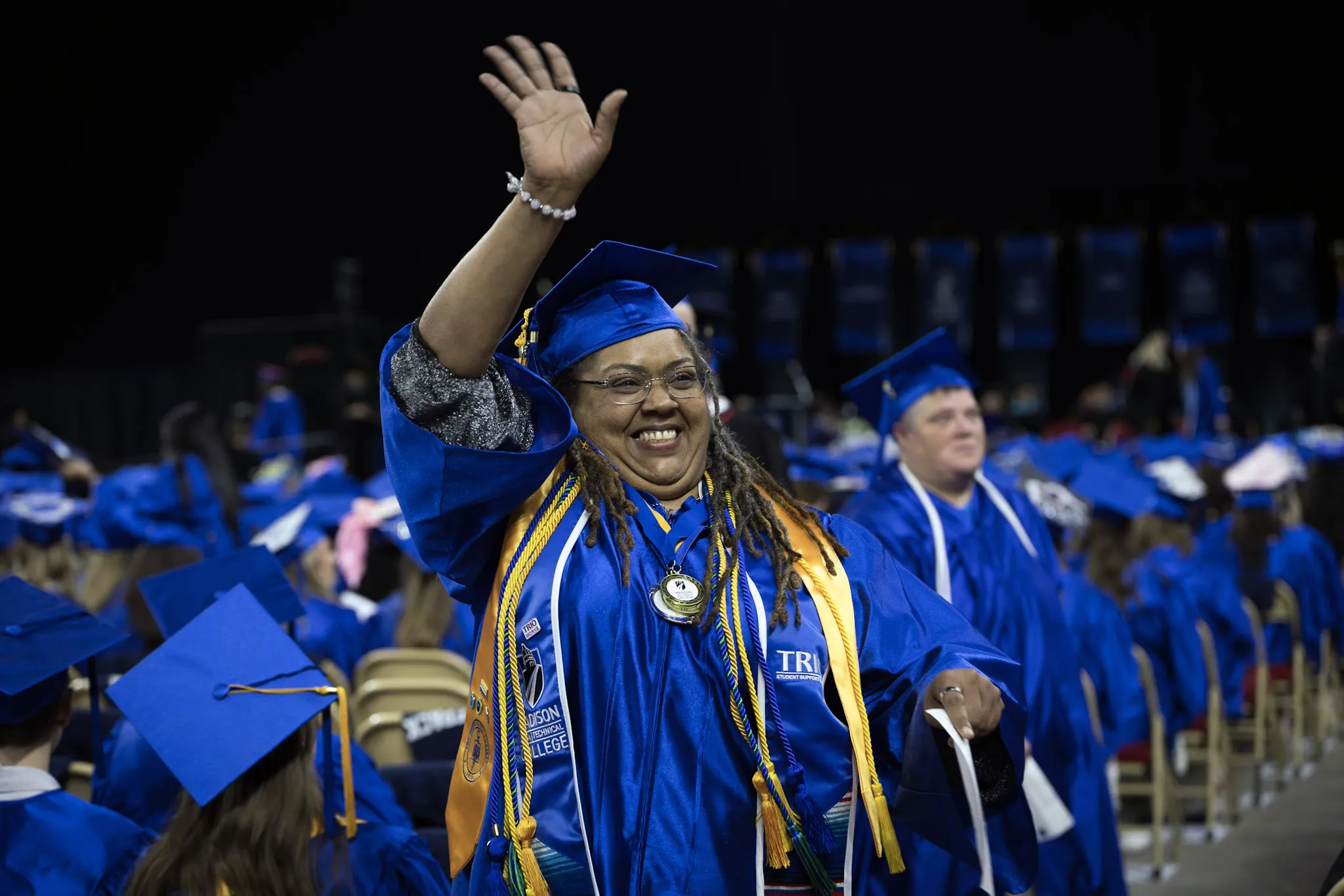 Woman grad with long black hair, wearing blue cap and gown, smiling and waving.