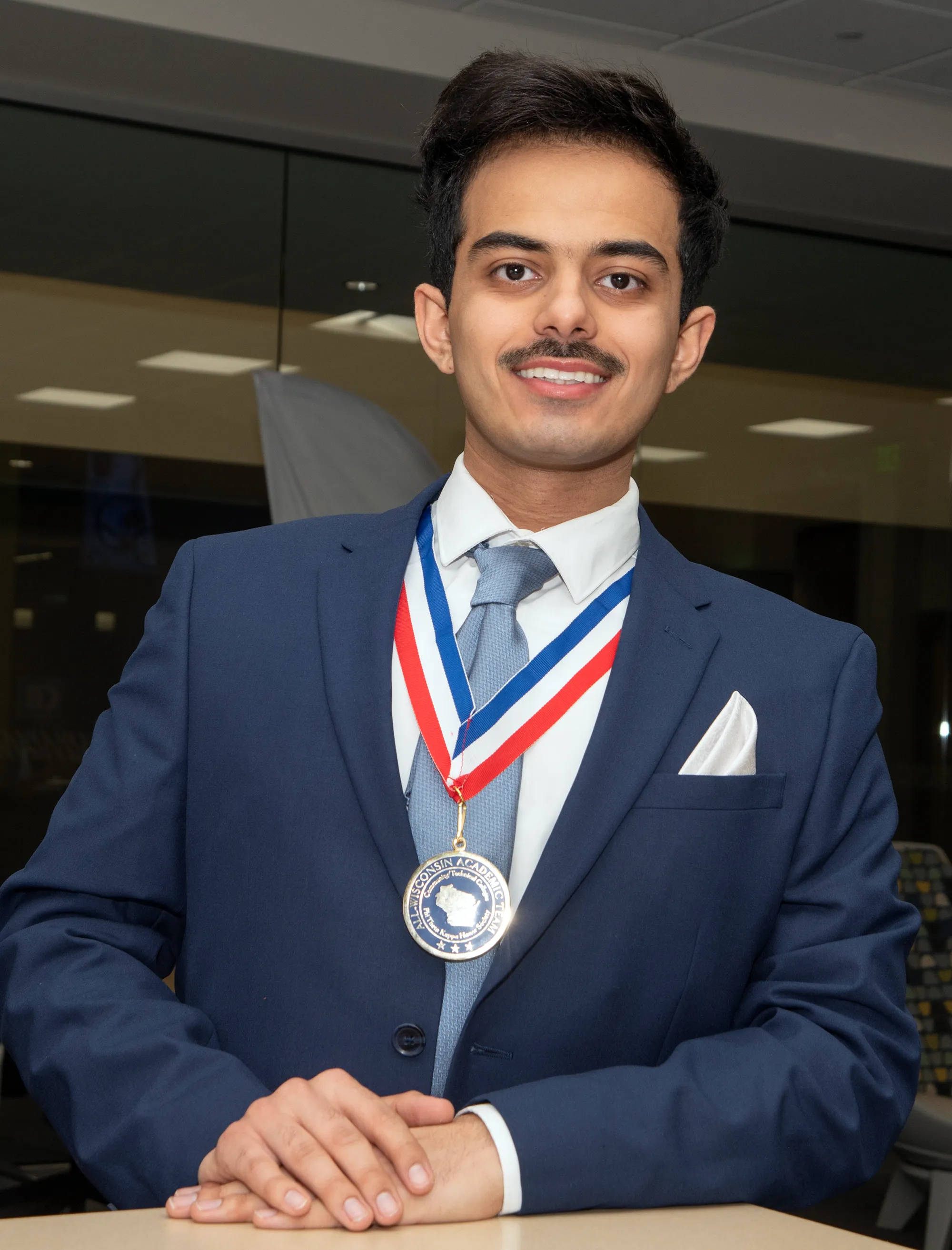 Student with dark hair, mustache, wearing dark blue blazer, with medal around his neck.