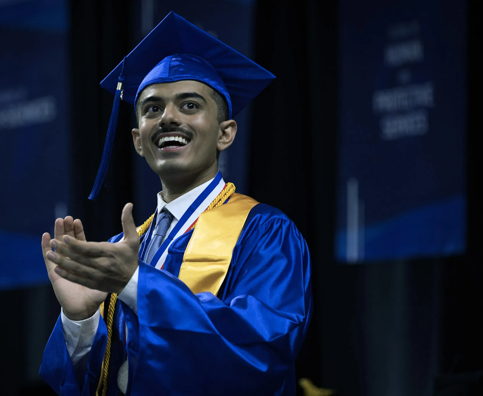 Man with blue cap and gown with yellow shawl, smiling and clapping.