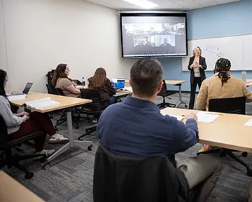 human resources classroom showing students sitting at tables while instructor lectures. Large screen in background showing hybird student attendees.