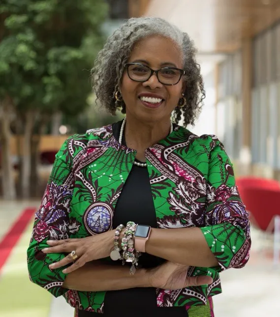 Person with grey short hair, black rimmed glasses, and multi-colored top, bracelets, smiling at camera.
