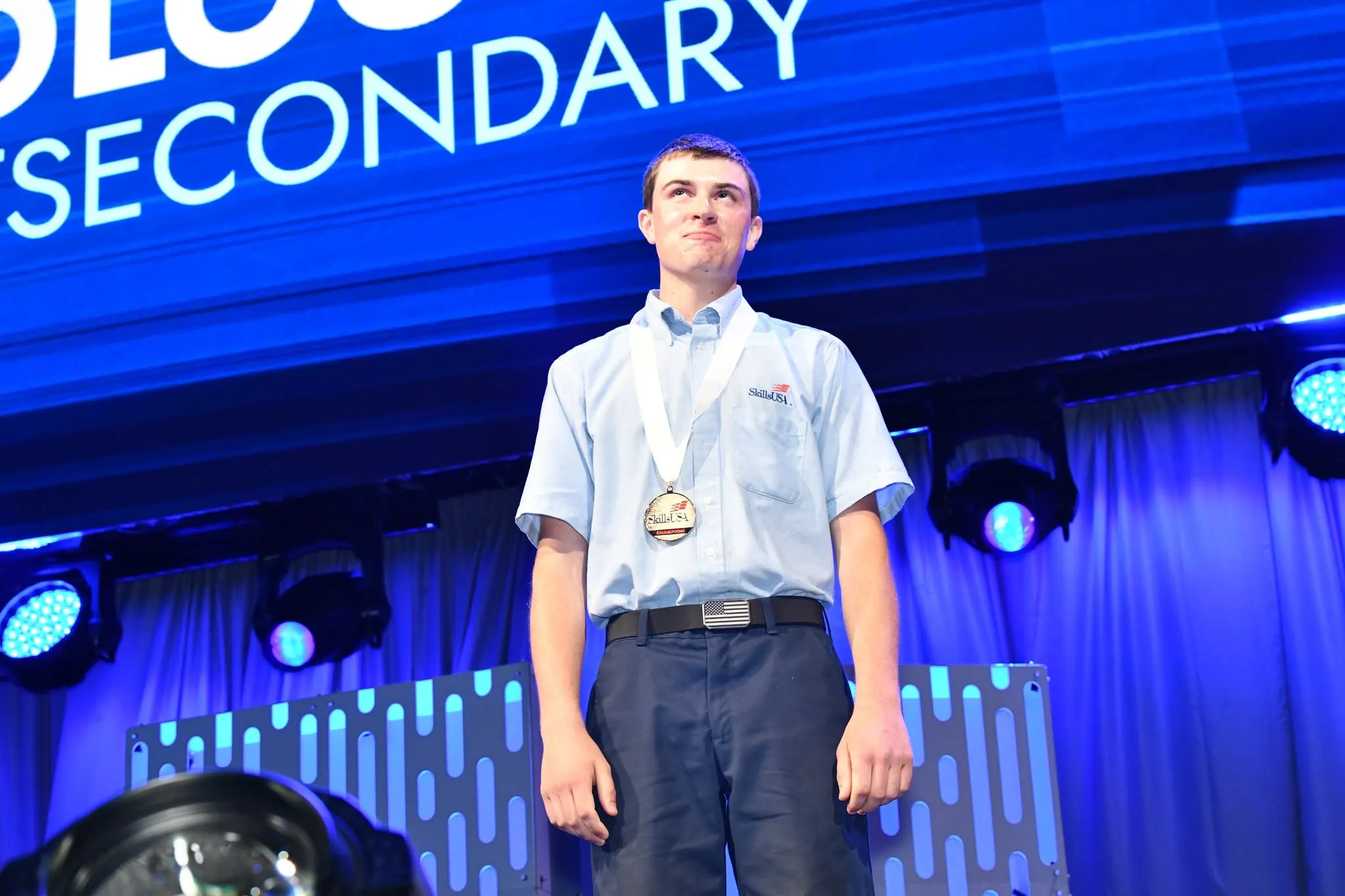 Male student with blue shirt and blue pants, wearing medal around neck, and smiling.