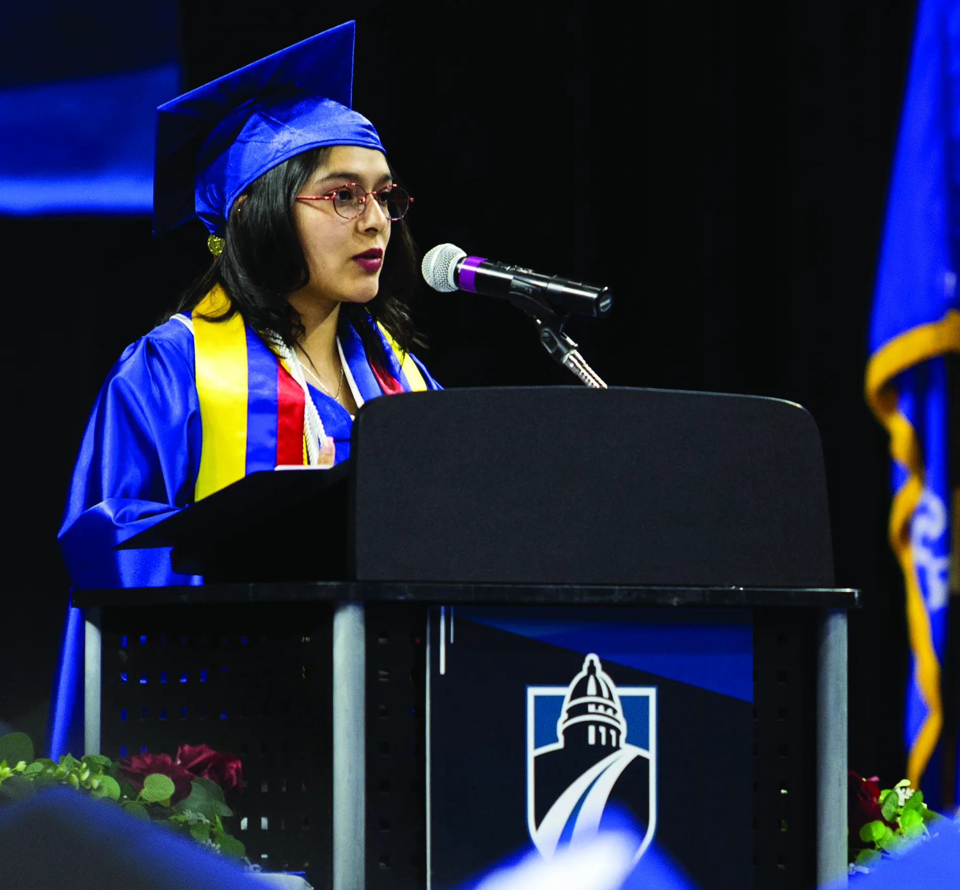 Female graduate at podium, speaking, wearing a blue cap and gown.