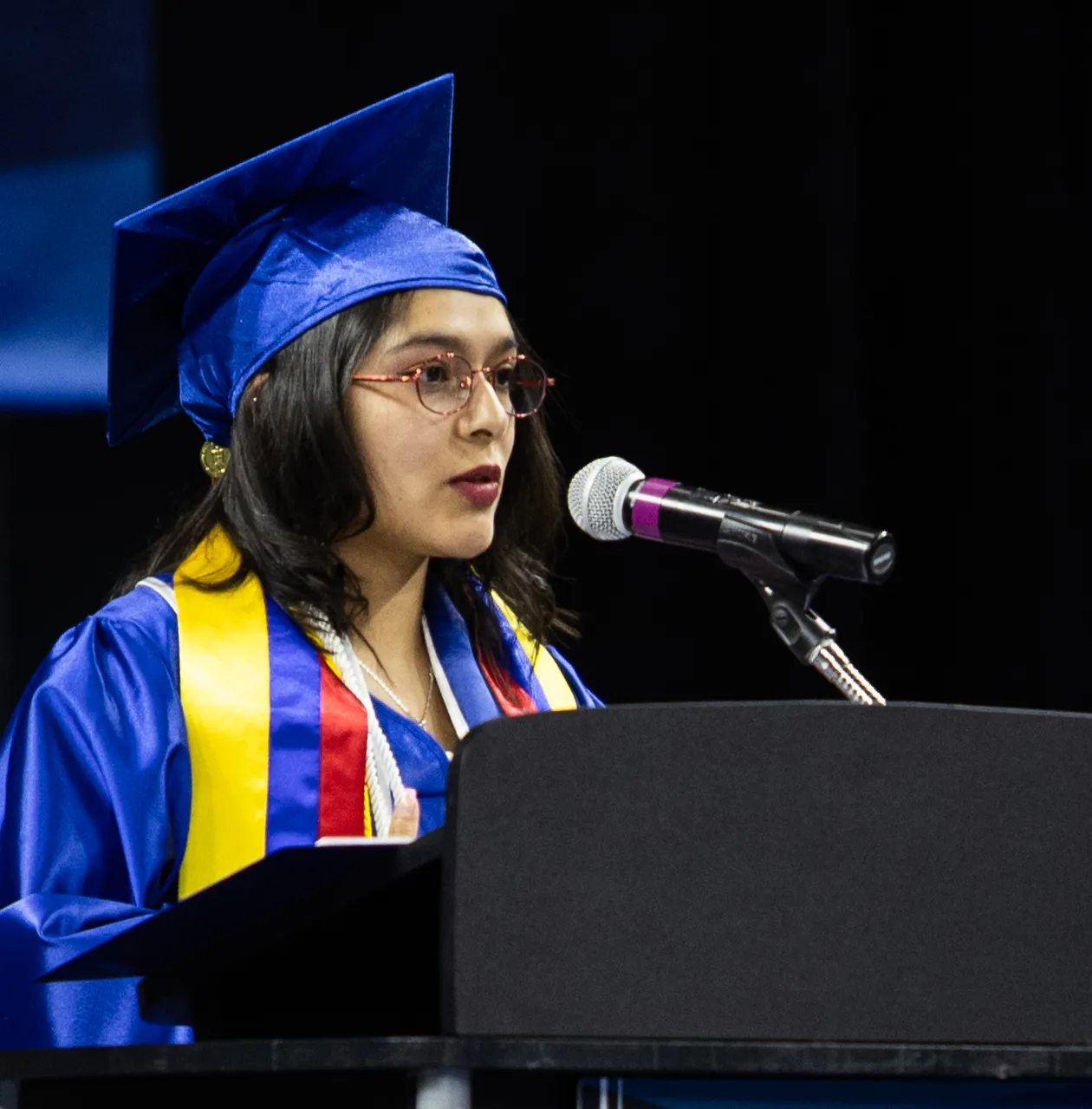 Grad at podium speaking, with blue cap and gown.