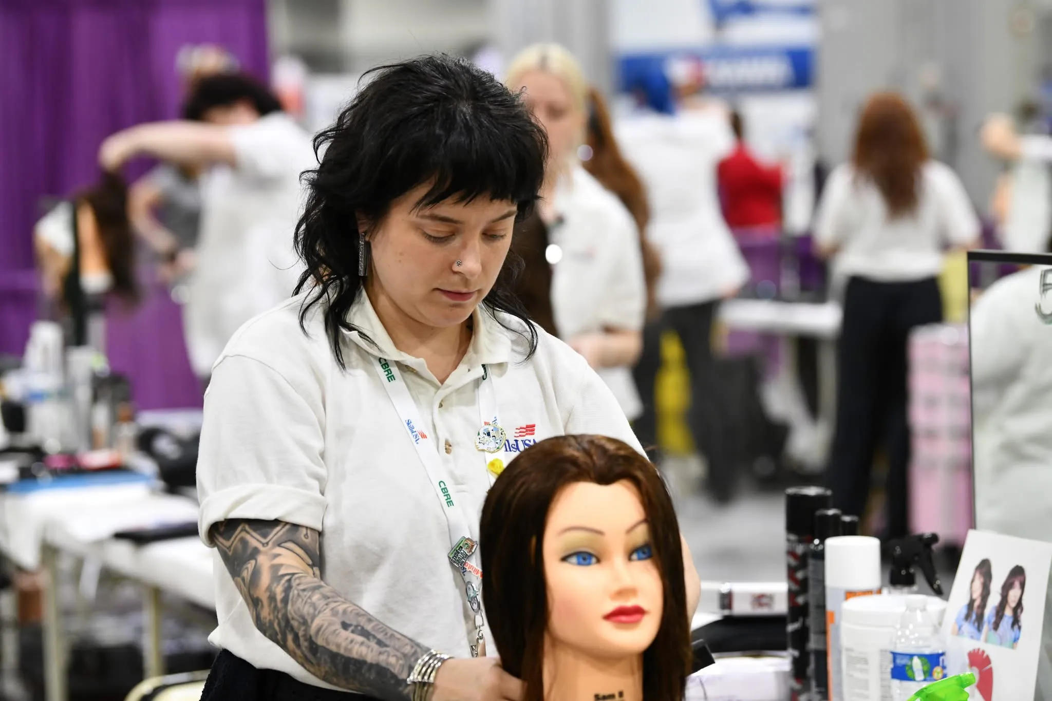 Student with black hair, white shirt, tattoos, styling the hair of a model dummy.