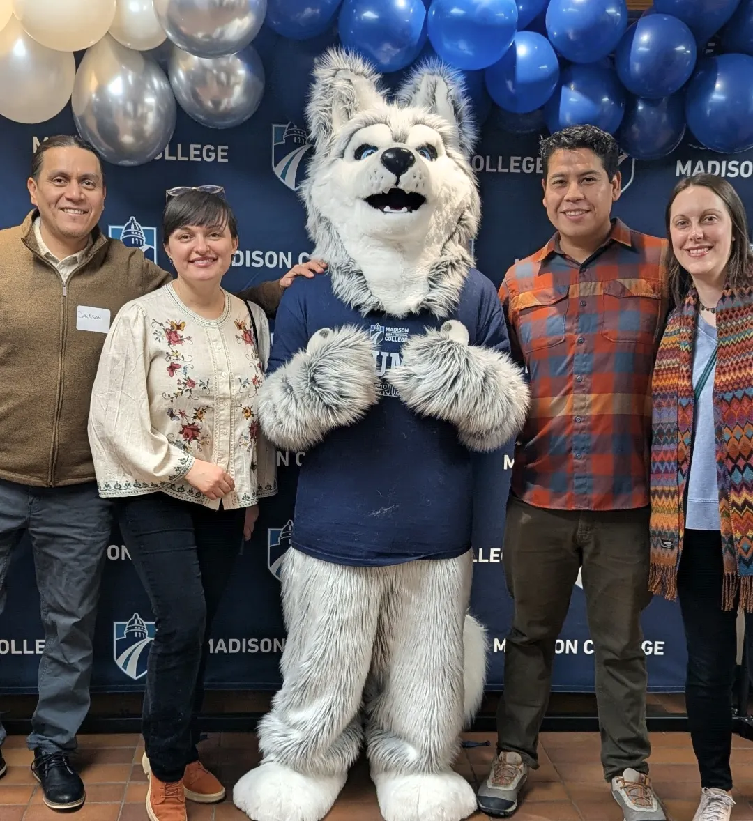 Four Madison College alumni posing with Wolfie mascot in front of a college logo banner.