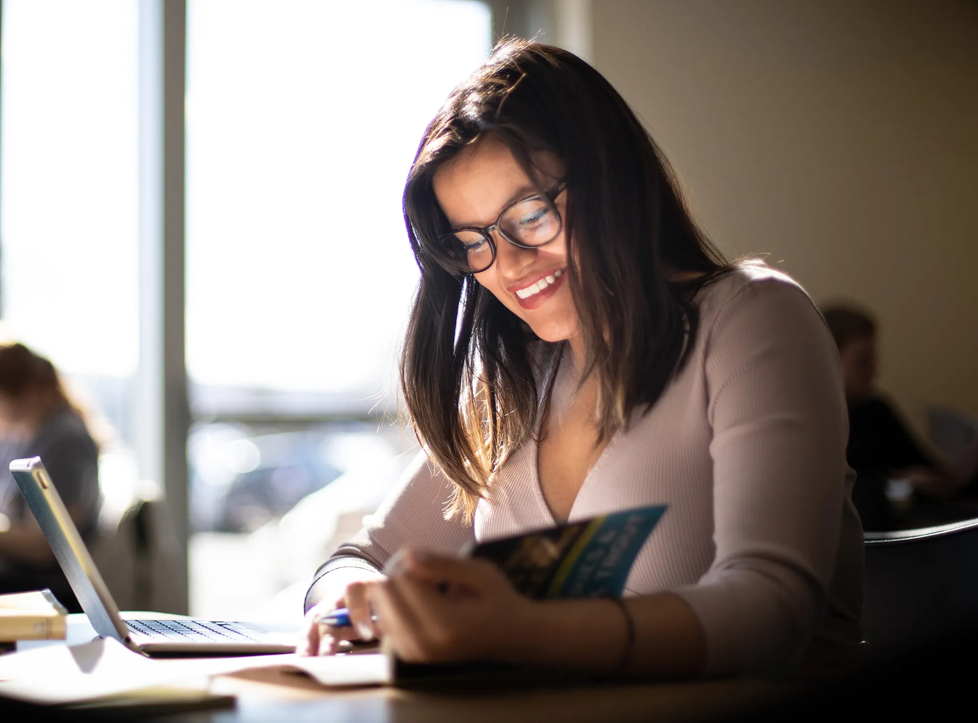Female student at desk, smiling and reading a book.