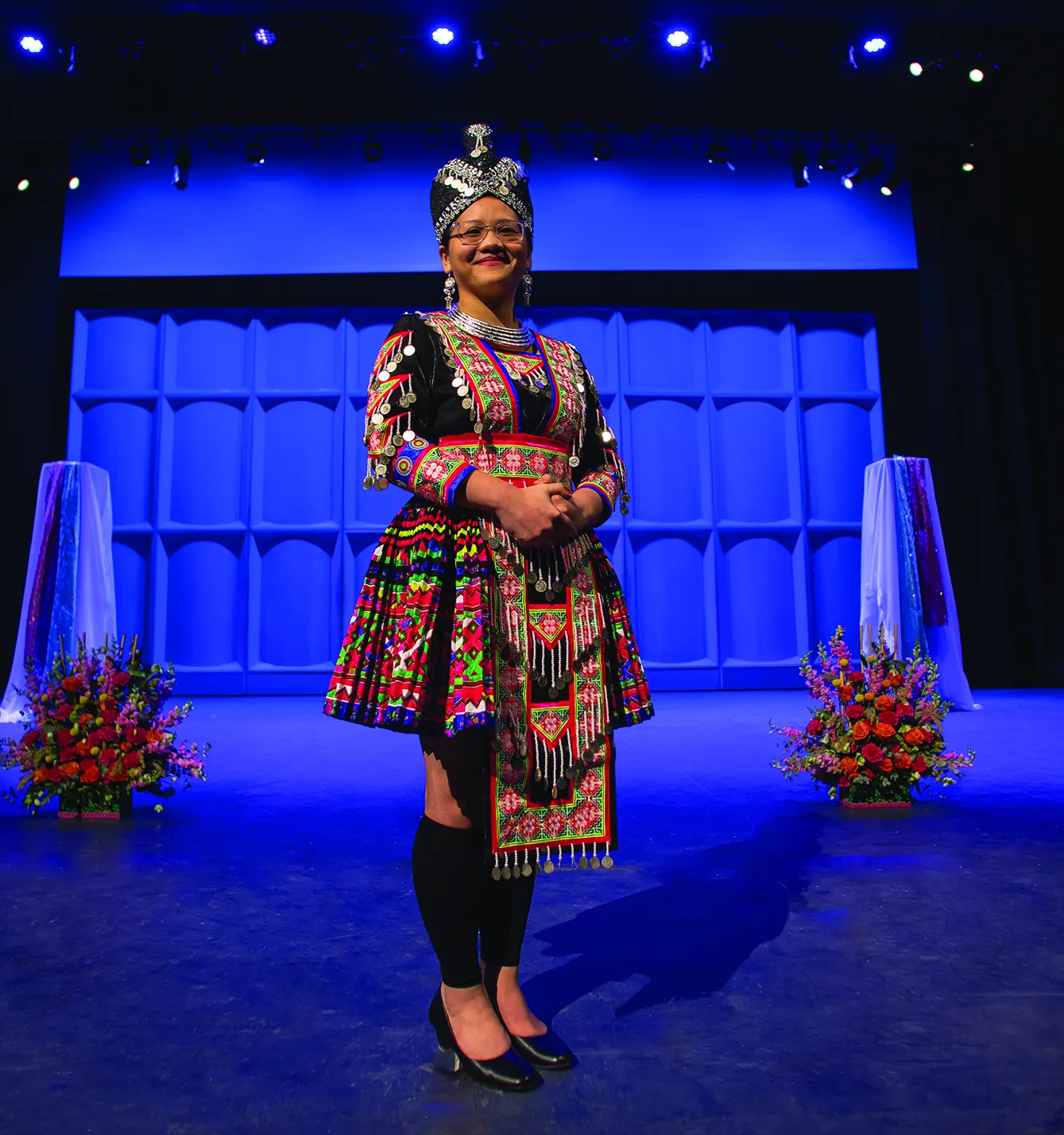 Women on stage wearing traditional Hmong clothing, a long multi-colored skirt, and headpiece.