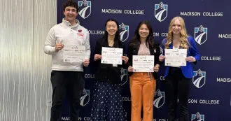 Four students, holding certificates, standing in front of a blue Madison College logo banner.
