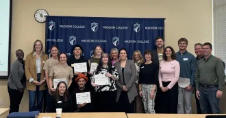 A group of people, posing in front of a blue Madison College banner.