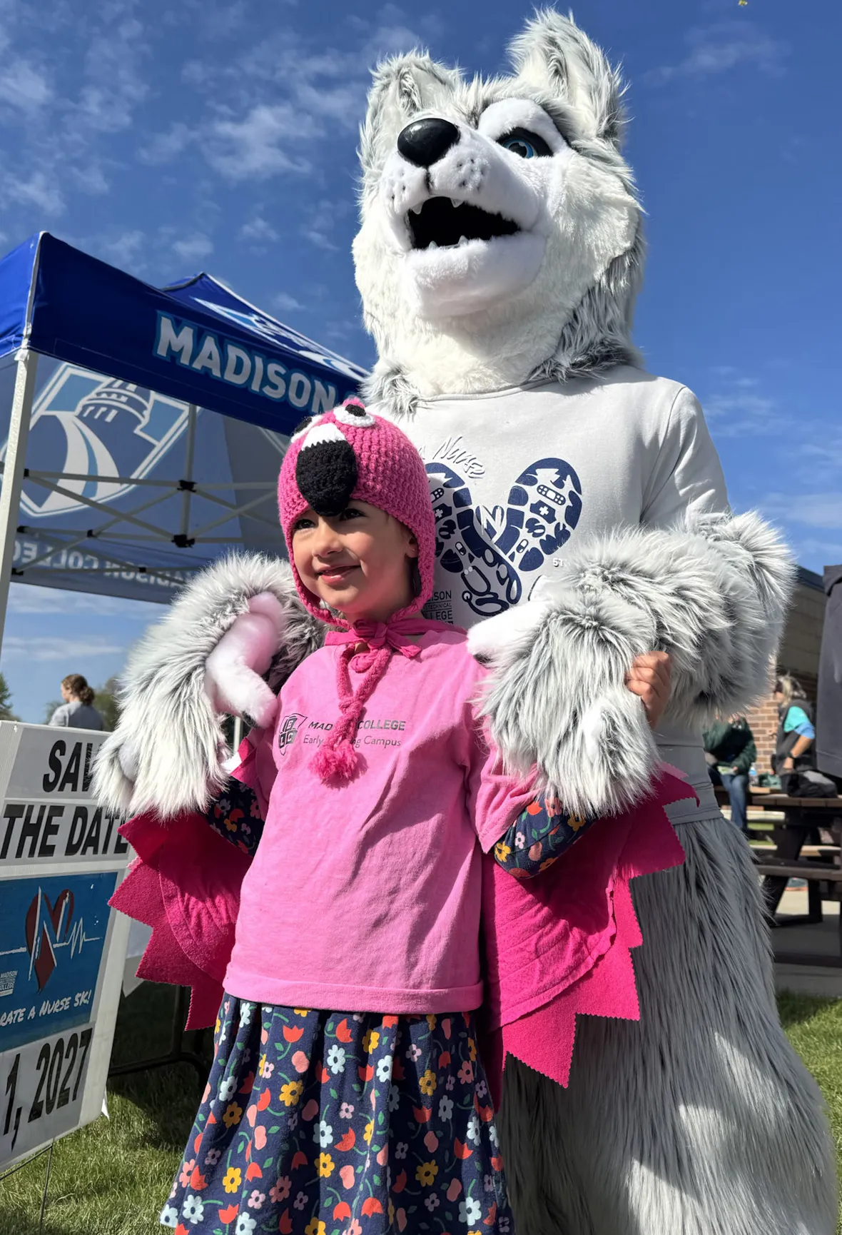 Wolfie mascot with grey fur and blue eyes, hugging a child with a pink shirt and flower patterned skirt.