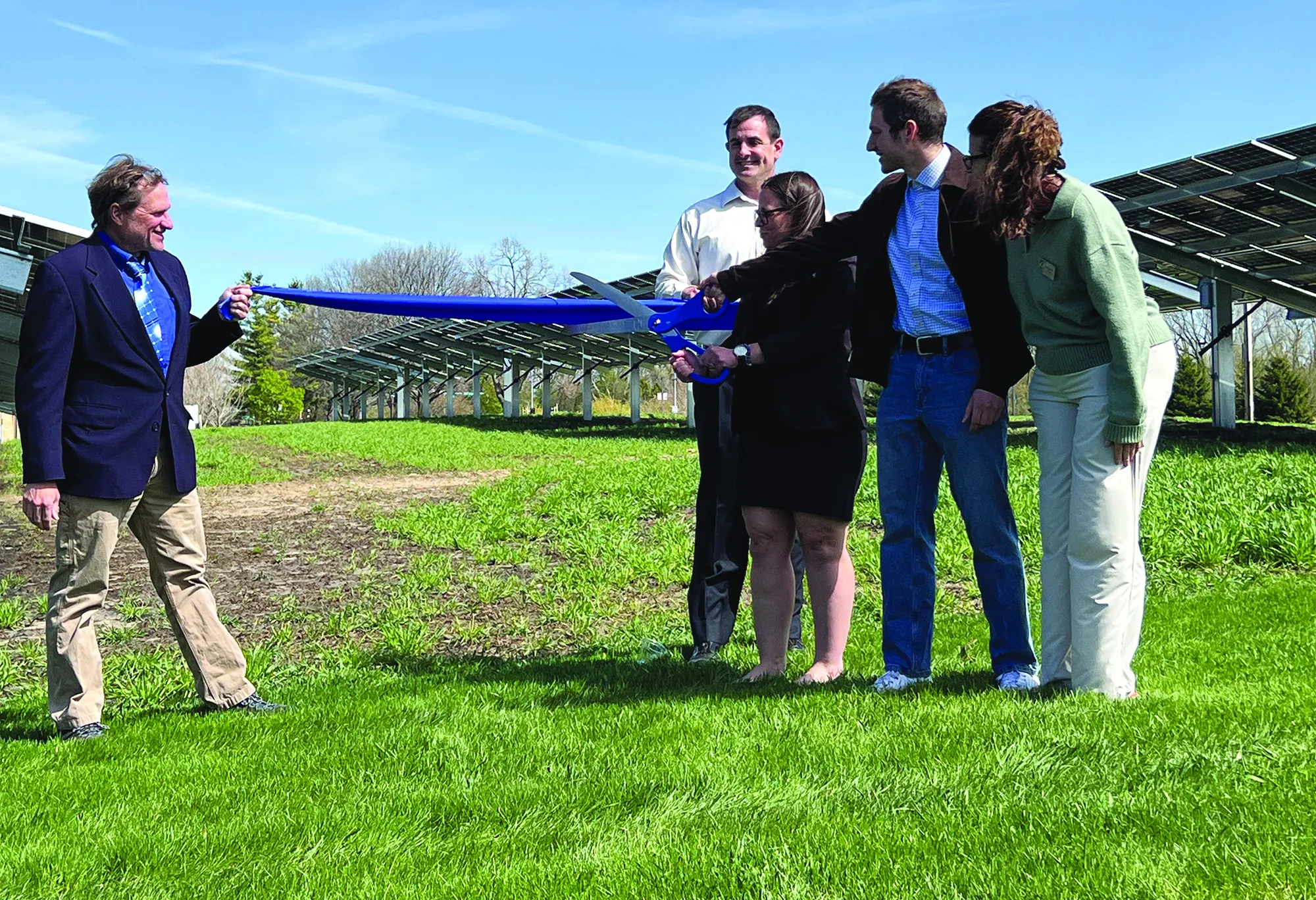Five Madison College faculty, student, leadership cut a blue ribbon as part of ceremony, outside with green grass, blue skies and solar panels in background.