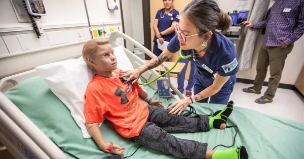 Students in a simulation clinic room holding a stethoscope to a simulated patient manikin boy, dressed in an orange shirt.