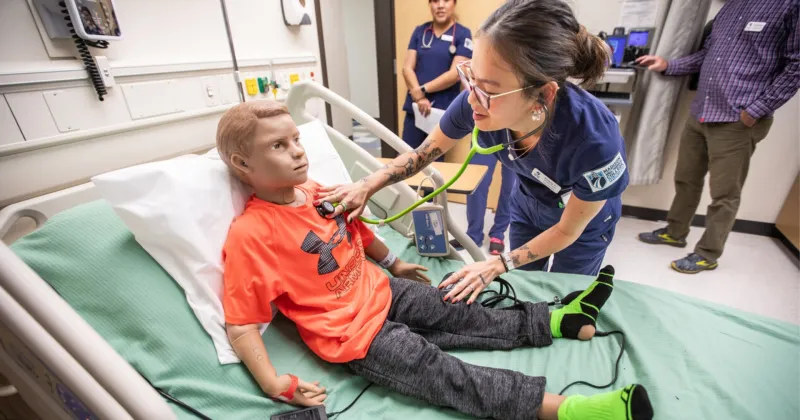Students in a simulation clinic room holding a stethoscope to a simulated patient manikin boy, dressed in an orange shirt.